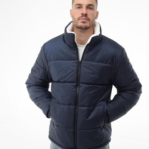 May include: A navy blue padded jacket with a high collar and a white, fluffy interior lining. The jacket features a full-length zip and pockets. The model is wearing the jacket with blue jeans.
