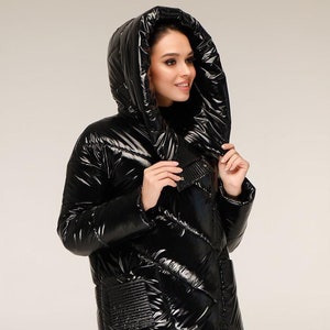 Può includere: Un lungo cappotto imbottito nero lucido con un ampio cappuccio. Il cappotto ha un design trapuntato e una chiusura a zip.