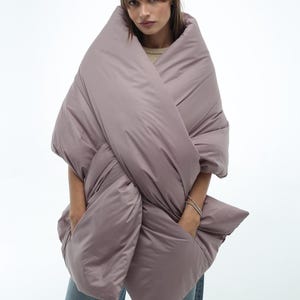 May include: An oversized puffer jacket in a mauve colour, worn as a wrap. The jacket is made from a soft, quilted fabric and has a relaxed fit.