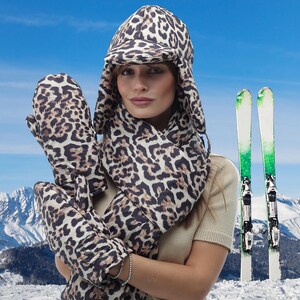 May include: A woman wearing a leopard print hat, scarf and mittens. The hat has ear flaps and a brim. The scarf is long and wraps around the neck. The mittens are fingerless and have a leopard print design. The woman is standing in front of a snowy mountain background.