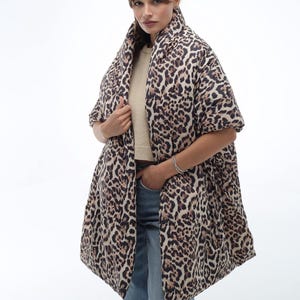 May include: A brown and black leopard print puffer scarf with a long, flowing design. The scarf is worn by a person with brown hair and is styled to drape over the shoulders and down the front of the body.