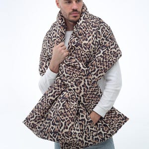May include: A leopard print scarf, with a pattern of brown and black spots on a beige background. The scarf is wrapped around the neck and shoulders. The person in the image is wearing a white long-sleeve shirt and blue jeans.