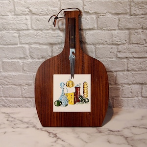 May include: A wooden cheese board with a decorative tile insert featuring a colorful illustration of food items. The board has a built-in cheese knife and a leather loop for hanging. The wood grain is visible.