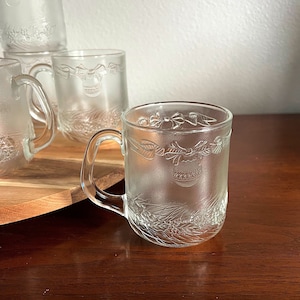 May include: Clear glass mugs with a frosted design. The mugs feature a festive embossed pattern of ornaments, bows, and foliage. The mugs have a handle and are suitable for hot or cold beverages. The mugs are displayed on a wooden surface.