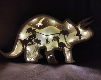 Könnte beinhalten: Eine weiße Holz-Dinosaurier-Nachtlampe mit einer Silhouette eines Triceratops, Palmen und anderen Dinosauriern. Das Licht wird mit warmweißen LED-Leuchten beleuchtet.