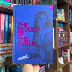 May include: A hardcover book titled "The Revolt of the Public" with a blue cover and pink lettering. The cover features an illustration of a woman and a smartphone. The book is held in front of a bookshelf.