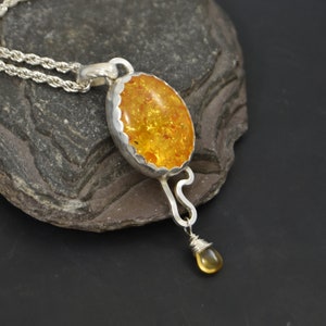 May include: Silver pendant necklace featuring an oval amber stone with a scalloped bezel. A smaller amber teardrop hangs below. The chain is a silver rope style. The pendant is displayed on a dark gray stone.