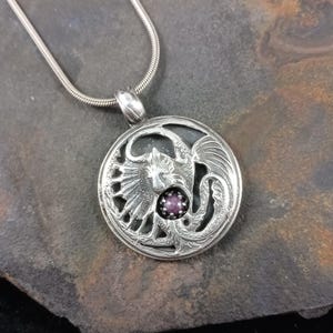 Könnte beinhalten: Silberne Halskette mit Anhänger, der ein detailliertes Drachenmotiv mit einem violetten Edelstein in der Mitte zeigt.