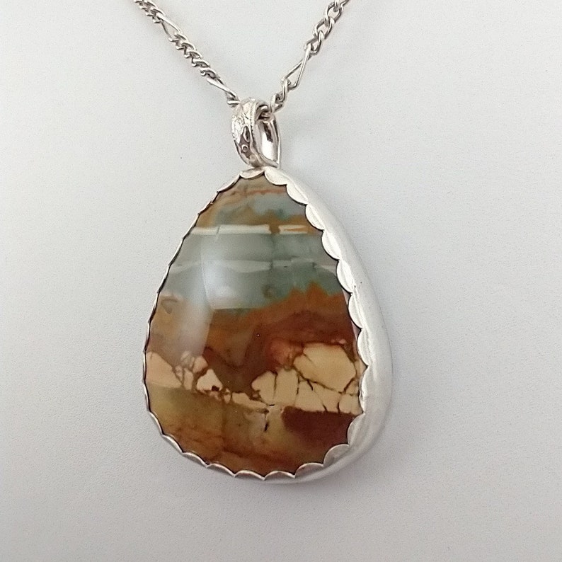 May include: A teardrop-shaped pendant necklace with a silver chain. The pendant features a polished stone with brown, beige, and blue-green hues, set in a scalloped silver bezel. The chain is a delicate, textured design.
