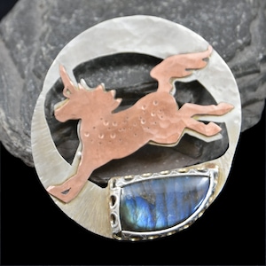 May include: A silver and copper unicorn pendant with a blue labradorite gemstone. The unicorn is leaping and the pendant is shaped like a crescent moon.