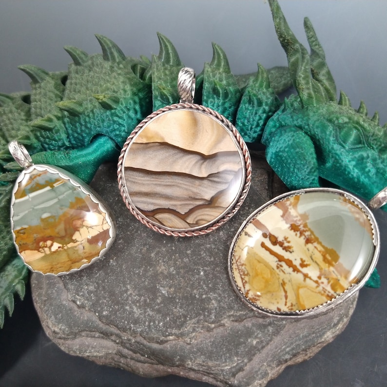 May include: Three gemstone pendants with silver-colored settings. The pendants feature unique patterns in shades of brown, green, and beige. One is round, one is oval, and one is teardrop-shaped. The pendants are displayed on a dark gray stone.