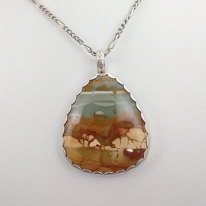 May include: A teardrop-shaped pendant necklace with a silver chain. The pendant features a large, polished stone with a mix of brown, beige, and blue-green colors. The stone is set in a silver bezel with a scalloped edge, creating a unique jewelry piece.