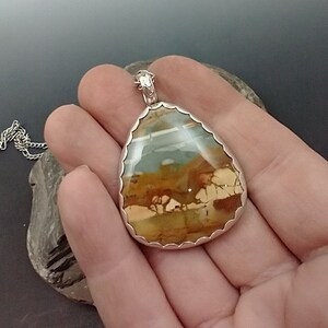May include: A teardrop-shaped pendant with a silver-colored scalloped frame. The pendant features a natural stone with a landscape-like pattern in shades of brown, beige, and blue-green. A silver chain is attached to the pendant.