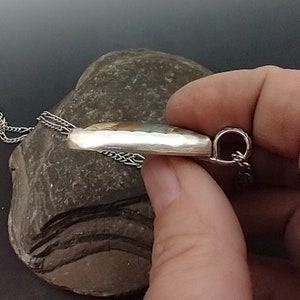 May include: A silver pendant with a hammered texture, featuring a colorful, oval-shaped stone. The pendant hangs from a delicate silver chain. The jewelry is displayed on a gray stone.