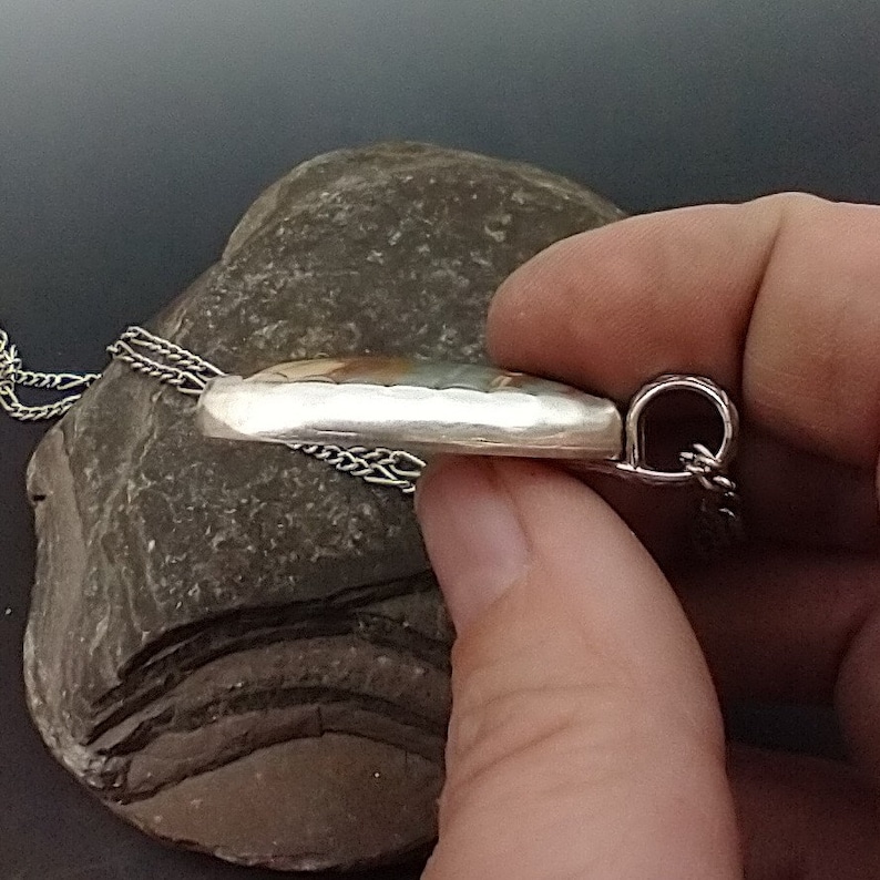 May include: A silver pendant with a hammered texture, featuring a colorful, oval-shaped stone. The pendant hangs from a delicate silver chain. The jewelry is displayed on a gray stone.