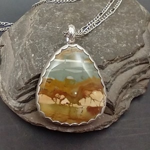 May include: A teardrop-shaped pendant with a silver-colored scalloped bezel and chain. The pendant features a polished stone with a landscape design in shades of brown, tan, and blue. The chain is a silver-colored, textured cable chain.