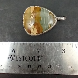 May include: A pendant with a unique, irregularly shaped stone, featuring a mix of brown, green, and cream colors. The stone is set in a silver-colored metal with a scalloped edge and a small loop for a chain. The pendant is approximately 2 inches long.