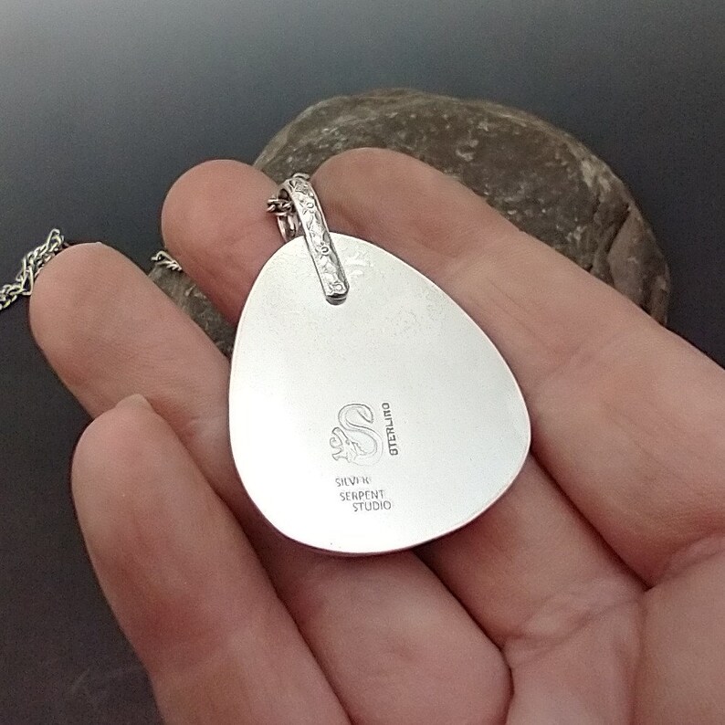 May include: A silver pendant with a teardrop shape, featuring a textured bail and a chain. The pendant's back is stamped with the words "STERLING" and "SILVER SERPENT STUDIO" around a stylized "S" logo. The pendant is held in a hand.