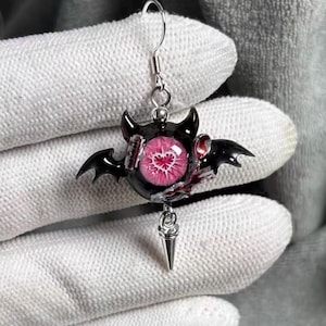 Peut inclure: Une boucle d'oreille pendante composée d'une sphère noire centrale avec de petites ailes de chauve-souris noires et un œil rose proéminent à pupille en forme de cœur. Elle comporte un crochet en métal argenté et une pointe en métal argenté.