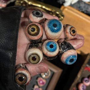 May include: A collection of rings featuring realistic, three-dimensional human eye designs. Each ring showcases a detailed iris in shades of blue, brown, or gray, surrounded by white sclera with visible red veins, all set on metallic bands.