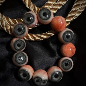 May include: A circular arrangement of spherical beads resembling human eyeballs with grey irises and black pupils, interspersed with smooth, flesh-toned, veined spherical beads, all resting upon a thick, twisted gold-coloured rope cord.
