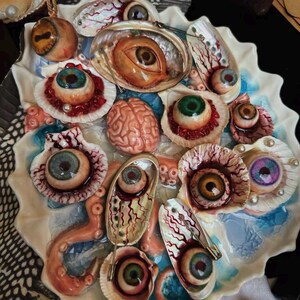 May include: A decorative tray featuring a collection of realistic sculpted eyeballs, a human brain model, and octopus tentacles nestled within various seashells. The arrangement includes iridescent abalone shells and white scallop shells with red veins.