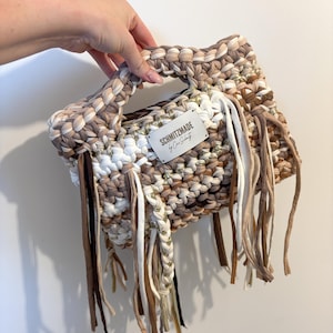 Puede incluir: Bolso de mano cuadrado hecho a mano en tonos beige, marrón y blanco. El bolso presenta un asa trenzada y flecos decorativos. Una pequeña etiqueta dice "SCHMITZMADE by Cris Schmitz".