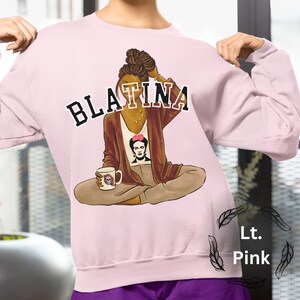 May include: A light pink sweatshirt with a graphic of a woman with brown skin and brown hair, wearing a brown cardigan and holding a coffee mug. The text "BLATINA" is printed in black letters above the woman.