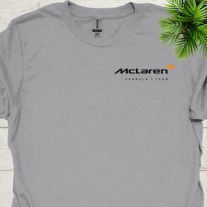 May include: A grey t-shirt with the McLaren Formula 1 Team logo in black and orange. The logo is on the chest, with the McLaren name above and "FORMULA 1 TEAM" below. The shirt has a crew neck and short sleeves.