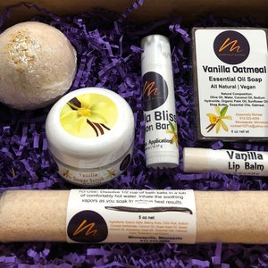 May include: A gift box filled with purple shredded paper and a variety of bath and body products, including a bath bomb, sugar scrub, lip balm, soap, and a lotion bar. The products are all vanilla scented and feature a vanilla flower design. The box is labeled "Essentially Michael" and includes the text "Vanilla Oatmeal Essential Oil Soap" and "Vanilla Lip Balm".