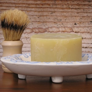 Op de afbeelding: Een witte keramische schaal met blauwe bloemenmotieven, met een ronde, lichtgele zeepstaaf en een bruin-wit scheerkwast.