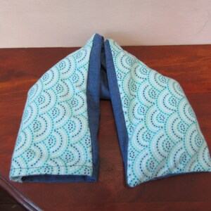 以下が含まれることがあります： 幾何学模様の入った青とターコイズのネックラップ。このラップは柔らかな生地で作られており、温熱パッドとして使用するために設計されています。