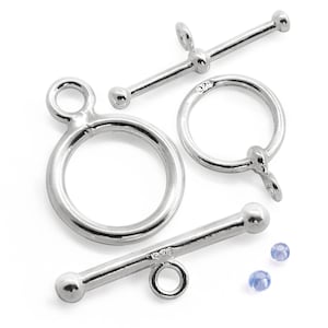 Pode incluir: Close-up de fechos de alternância prateados e duas pequenas contas azuis. Os fechos incluem anéis circulares e barras com extremidades em forma de bola. Os componentes estão dispostos sobre um fundo branco, ideais para a fabricação de joias.