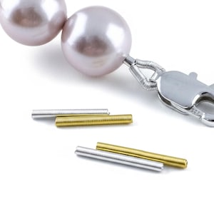 May include: A string of large, smooth, pale pink pearls attached to a silver lobster claw clasp, accompanied by four small, cylindrical metal crimp covers in alternating silver and gold tones, arranged in two pairs on a plain white surface.