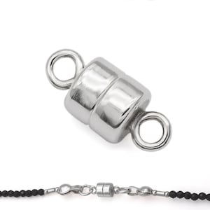 May include: Silver magnetic clasp for necklaces and bracelets. The clasp is made of two cylindrical pieces that connect with magnets. The clasp is shown in a close-up view and in a full view on a black beaded necklace.