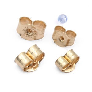 May include: A collection of gold-colored earring backs and a small, light blue bead. The earring backs are in various shapes, including butterfly and barrel styles. The items are arranged on a white surface, showcasing jewelry components.