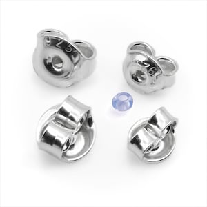 Puede incluir: Cuatro cierres de pendientes plateados y una pequeña gema azul claro están dispuestos sobre una superficie blanca. Los cierres tienen forma redondeada con un orificio para el poste y un diseño curvo y seguro. La gema es redonda y translúcida.