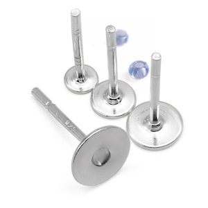 Può includere: Primo piano di quattro perni per orecchini color argento con basi piatte e rotonde. I perni sono disposti su una superficie bianca, con due piccole perle blu sullo sfondo. I perni degli orecchini hanno un diametro di circa 0,6 cm.