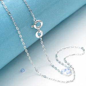 May include: A delicate silver necklace with a small, oval-link chain. The necklace features a spring ring clasp and a small, round tag. The background is a light blue color.