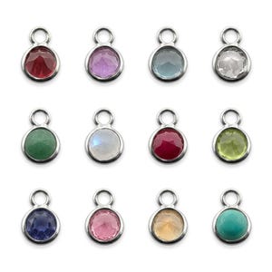 Op de afbeelding: Een verzameling van twaalf ronde metalen bedels, elk voorzien van een gefacetteerde edelsteen in diverse kleuren, waaronder rood, paars, lichtblauw, transparant, groen, iriserend wit, donkerrood, limoengroen, donkerblauw, roze, amber en turkoois.