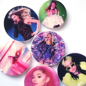 以下が含まれることがあります： 異なる女性歌手の画像が印刷された、丸い形の携帯電話グリップが6つ。グリップは光沢のある白い仕上げです。歌手は、アリアナ・グランデ、ビリー・アイリッシュ、テイラー・スウィフト、そして金髪の歌手の異なる画像が3つです。