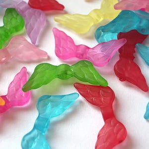 Puede incluir: Una colección de coloridos amuletos de cola de sirena translúcidos en varios tonos de rosa, azul, verde, rojo y amarillo. Estos amuletos decorativos están dispersos sobre una superficie blanca, mostrando sus diseños detallados en forma de aleta. Ideal para manualidades o joyería.