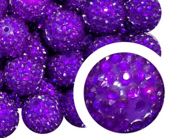 May include: A collection of large, round, purple beads with a textured, faceted surface. A magnified view of a single bead is shown in the lower right corner. The beads have a sparkly, glittery appearance. The beads are clustered together.