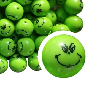 Pode incluir: Contas redondas verde brilhante com rostos de desenhos animados pretos. As contas têm um acabamento brilhante e um furo para enfiar. A imagem inclui um close-up de uma conta, mostrando os detalhes do rosto. Estas contas são adequadas para fazer joias e artesanato.