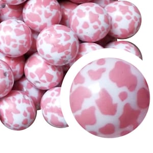 以下が含まれることがあります： ピンクの牛柄模様の白い丸いビーズのコレクション。ビーズは滑らかで光沢があり、右下にビーズのクローズアップがあります。ジュエリー作りや工芸品に適しています。