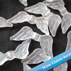 May include: Clear, translucent angel wing charms scattered on a dark surface. The wings have detailed feather textures and are slightly curved. A blue banner in the corner reads "More Colors Available!"