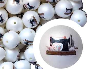 May include: White round beads with a sewing machine design. The beads have a glossy finish and a small hole for stringing. The sewing machine illustration is black, red, and brown. These beads are suitable for jewelry making and craft projects.