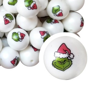 May include: White round beads with a cartoon image of a green character wearing a red Santa hat. The beads are suitable for crafting and jewelry making. The Grinch-themed beads are a festive addition to holiday projects.