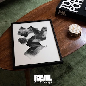May include: A black and white abstract painting in a black frame on a wooden table. The painting is of a geometric design with black brushstrokes on a white background.