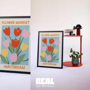 May include: A black framed print featuring a colourful illustration of flowers with the text "FLOWER MARKET AMSTERDAM". The print is displayed on a red shelf with a small plant and other decorative items.